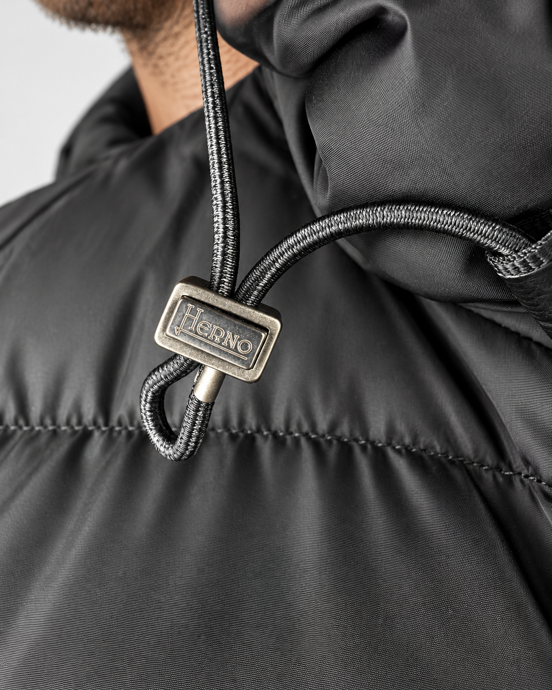 Dettaglio della coulisse del cappuccio con fermacorda metallico firmato Herno sul piumino nero.